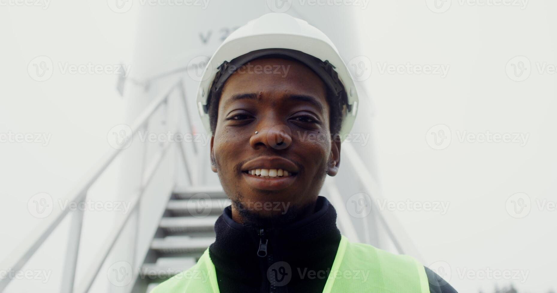 African American man in a green vest and hard hat smiles with his teeth looking at camera, standing near the entrance to an industrial wind turbine, close up, dynamic . in 4k, red komodo photo