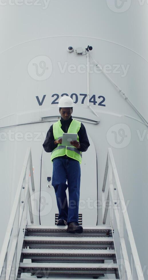 A young African American man wearing a white hard hat and green vest uses a tablet while walking down the steps of a wind turbine during an inspection. in 4k, red komodo photo