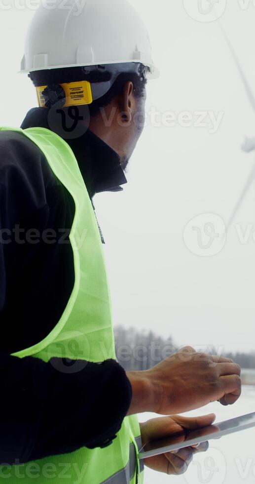 African American man in a work uniform with a hard hat on his head uses a tablet while standing near a wind turbine and looking at it, winter day, close up, vertical . in 4k, red komodo photo
