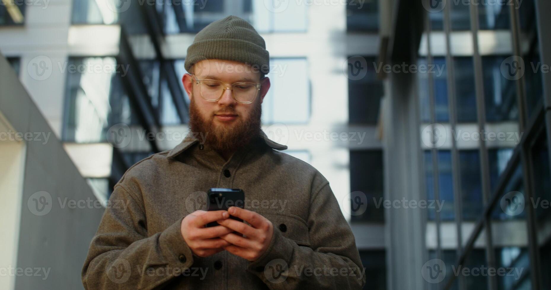 A young man in a knitted hat with a voluminous red beard is typing on a mobile phone while walking around the business center of the city, close up. in 4k, red komodo photo
