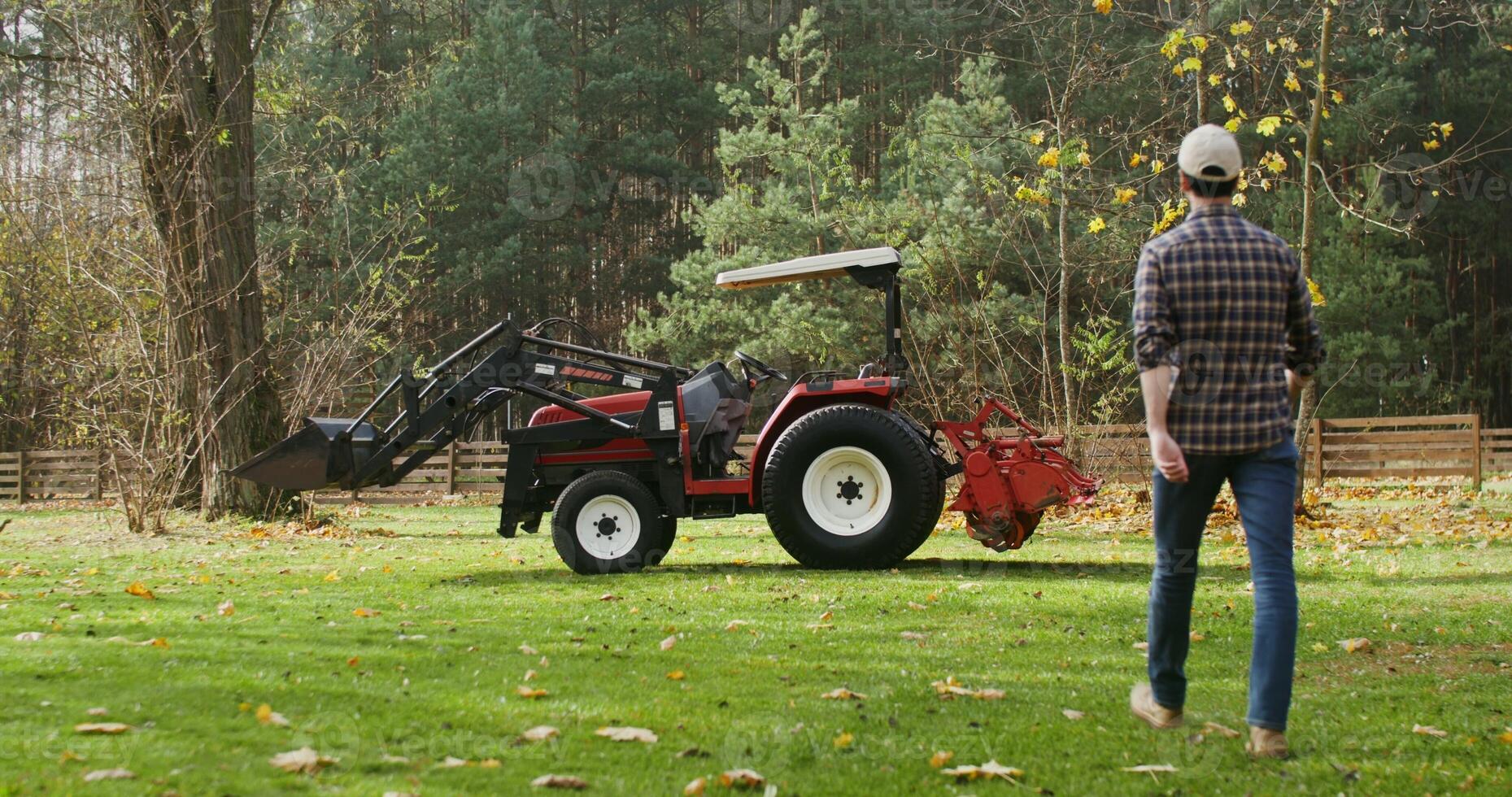 A young dark haired man in a baseball cap walks up to a modern tractor parked at the edge of a farm and gets into it, getting ready to work in a field on a sunny autumn day photo