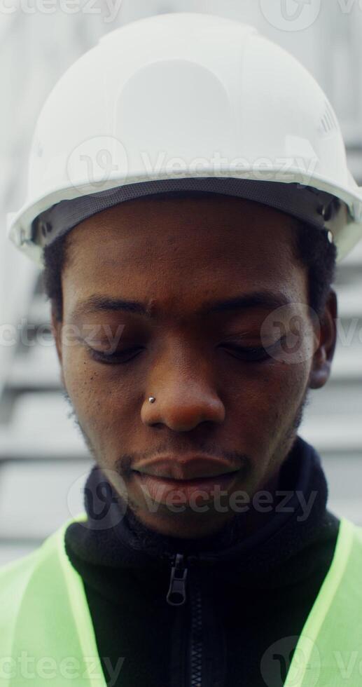 A young African American man in a white hard hat and a green vest looks at the camera with a smile, raising his head, close up. in 4k, red komodo photo