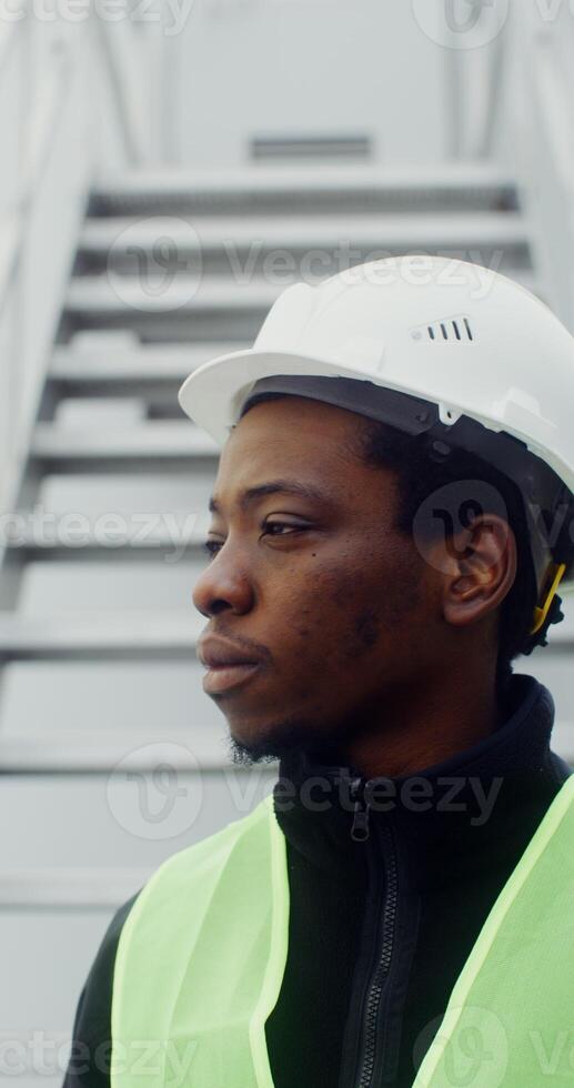 A young African American man in a white hard hat and green vest turns his head to look at the camera and smiles, standing near the entrance to the wind turbine, vertical . in 4k, red komodo photo