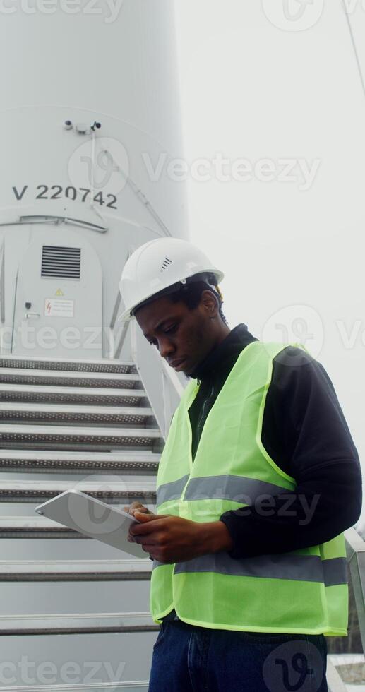 A young African American man in a green vest and hard hat uses a tablet while standing near the entrance to an industrial wind turbine, vertical . in 4k, red komodo photo