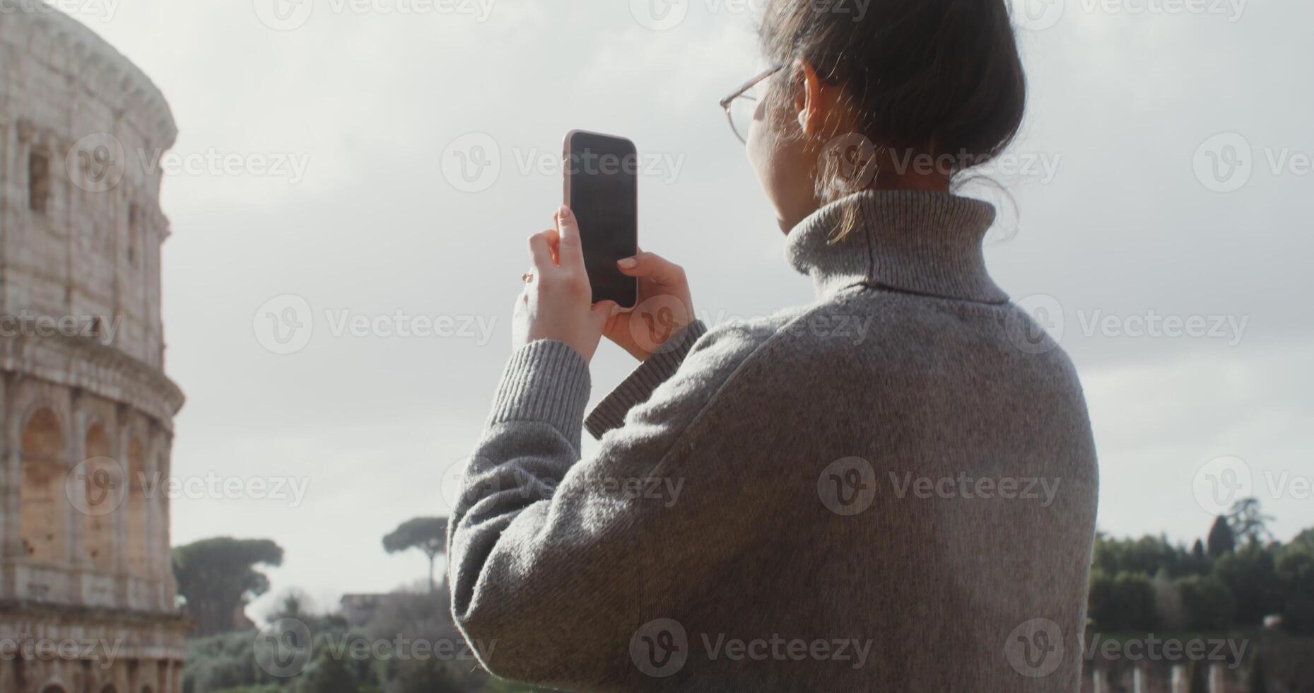 A young woman of European appearance in glasses, dressed in a warm sweater, photographs the Colosseum on a mobile phone, close up. Italy Rome. in 4k, red komodo. photo