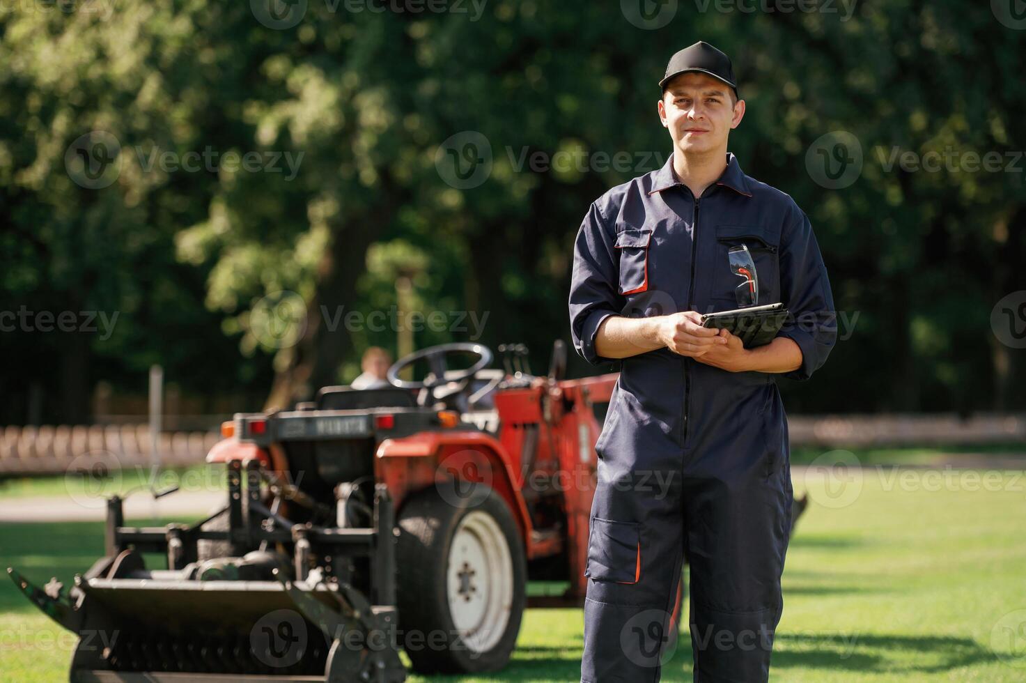 In ,,cap, holding digital tablet., Man is with utility tractor with grass cutter and aerator equipment on the field photo