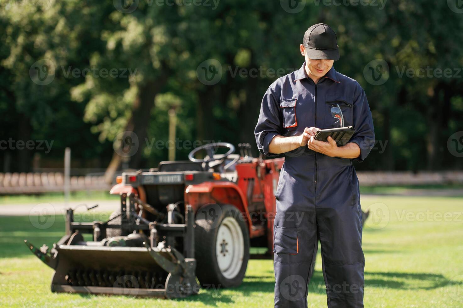 In ,cap, holding ,digital,, tablet. Man is with utility tractor with grass cutter and aerator equipment on the field photo
