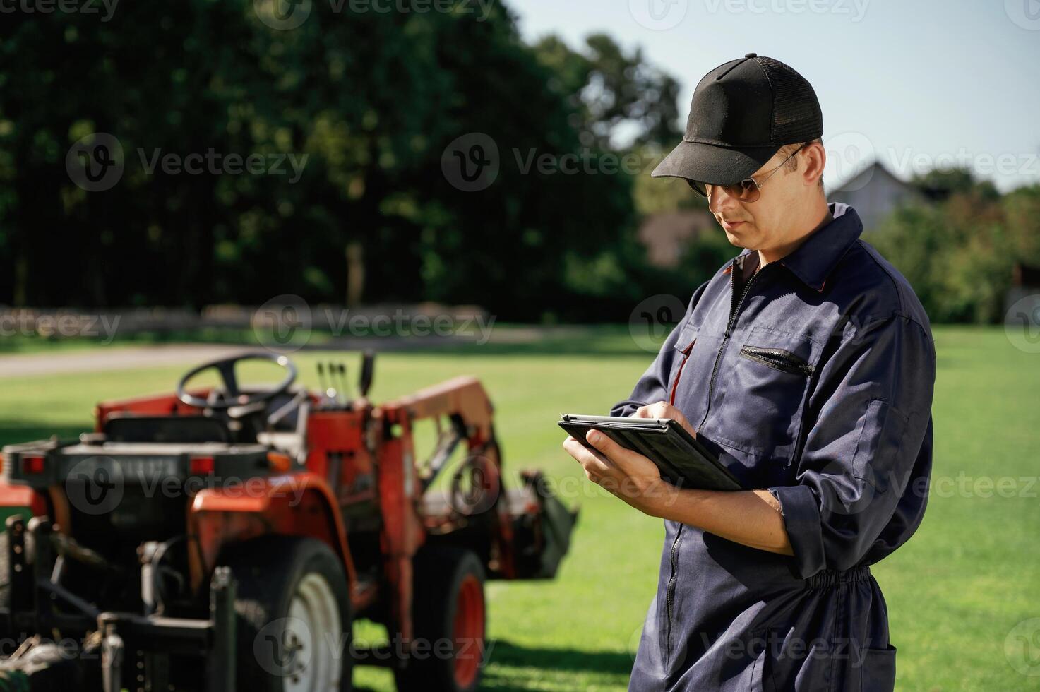 Black digital tablet in ,hands. Man is with utility tractor with grass cutter and aerator equipment on the field photo