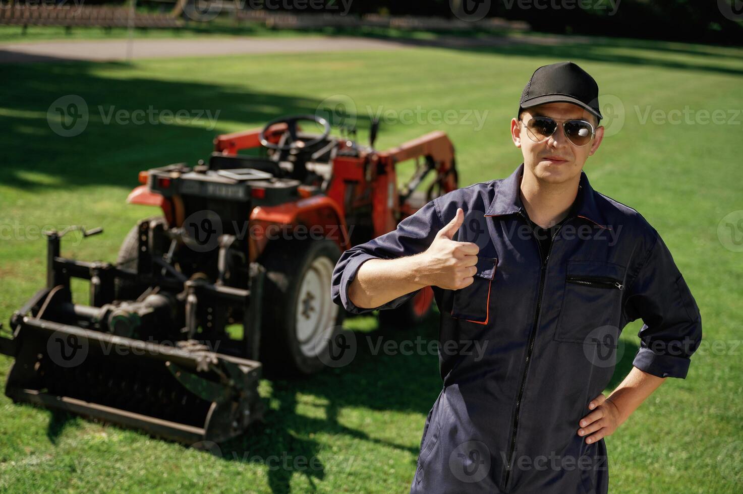 Shows, thumb up. Man is with utility tractor with grass cutter and aerator equipment on the field photo