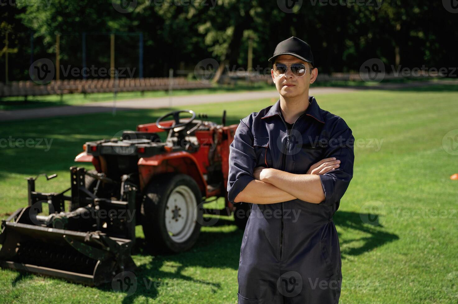 Standing ,with arms crossed. Man is with utility tractor with grass cutter and aerator equipment on the field photo