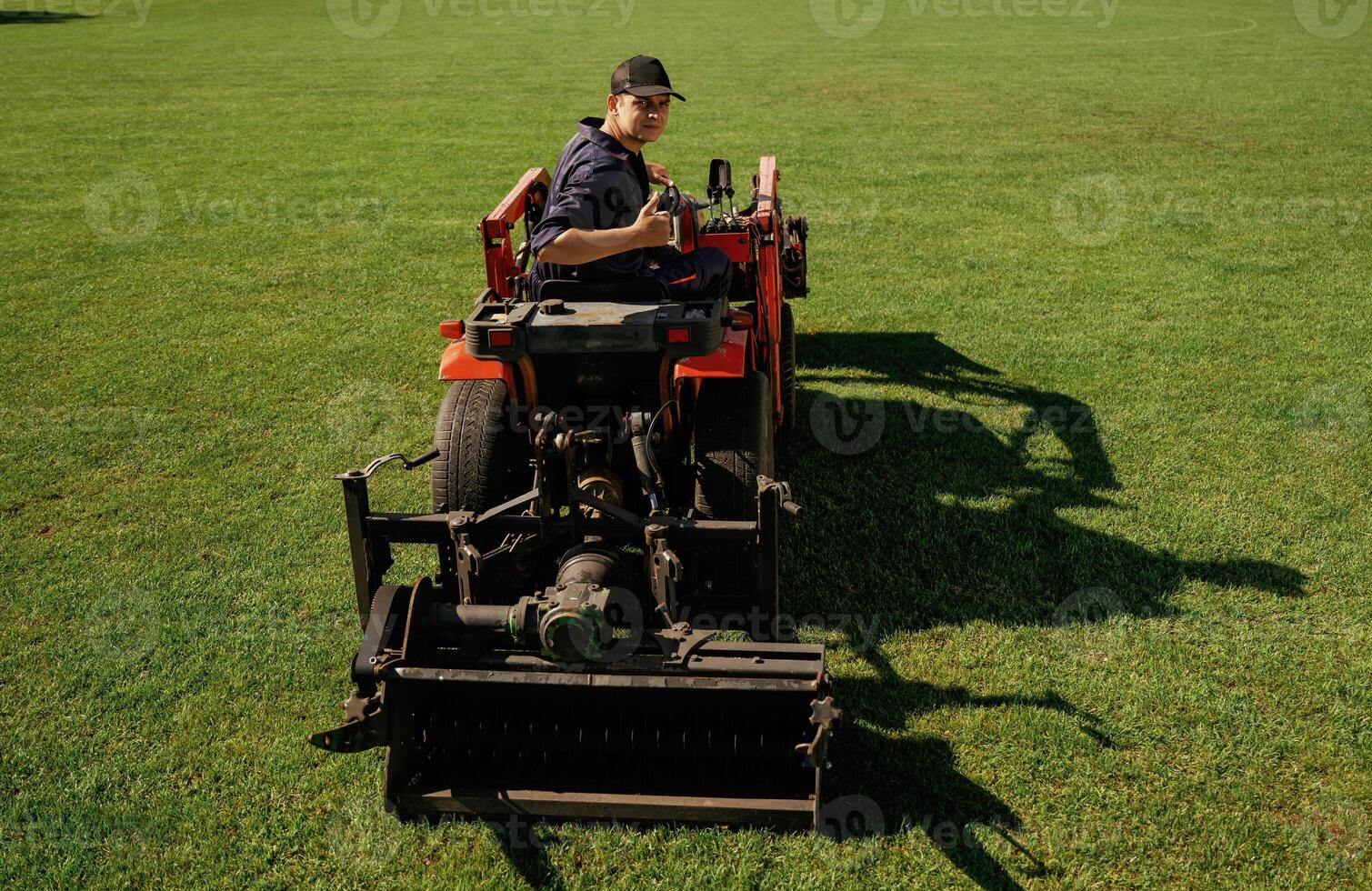 Driving, looks, behind, shows thumb up. Man is with utility tractor with grass cutter and aerator equipment on the field photo