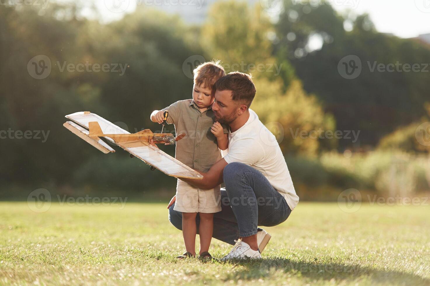 Trying ,to fix it with screwdriver. Father ,,and son are on the field together with toy plane photo