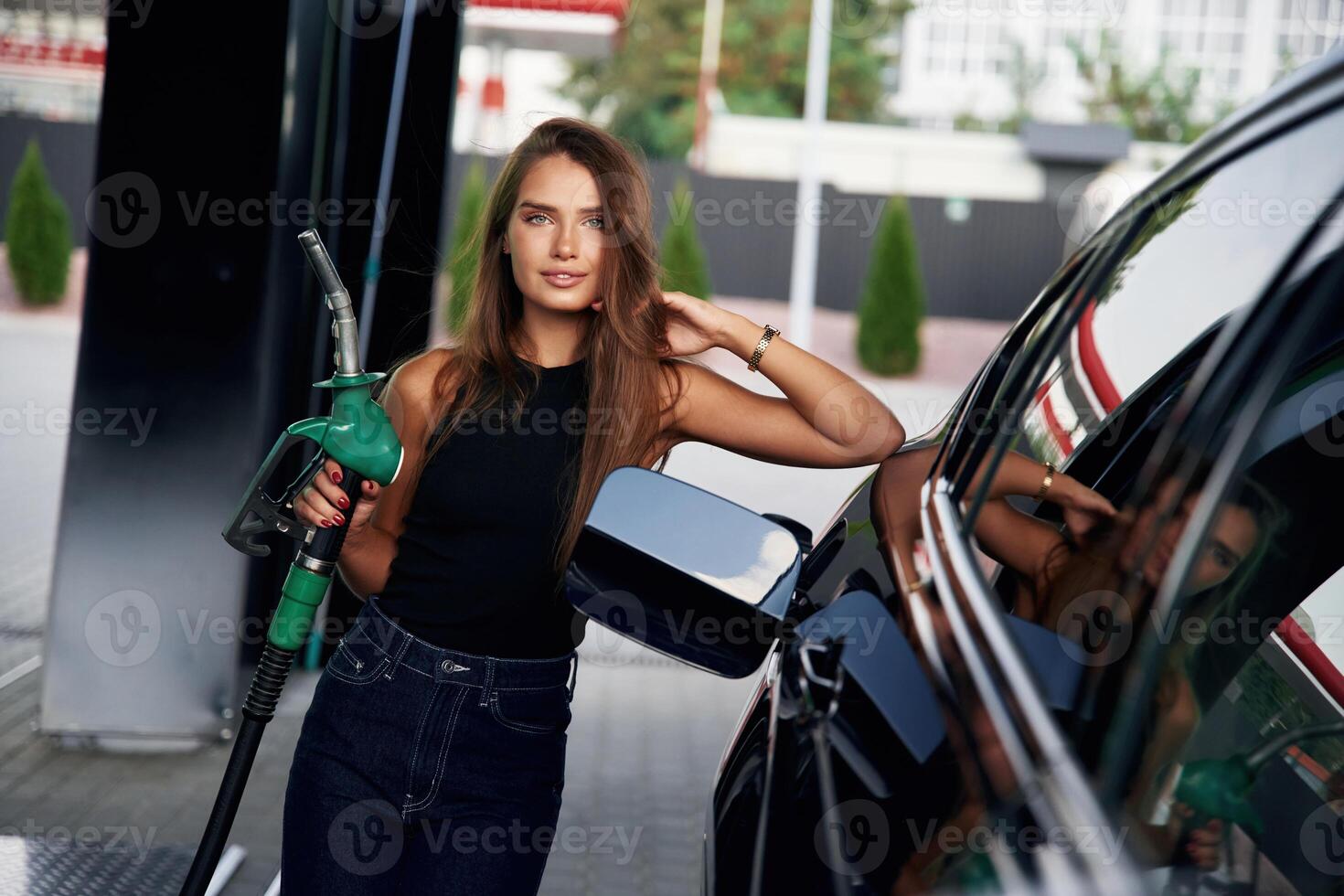 Leaning on, the, automobile, holding pump nozzle. A young woman at a gas station with her car photo
