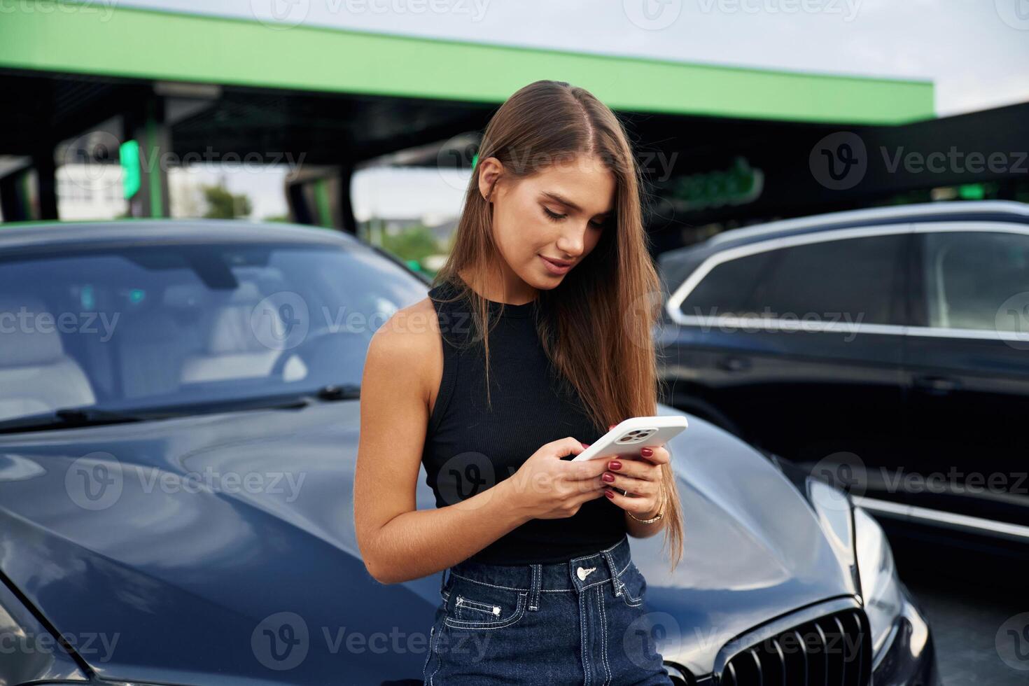 Scrolling the internet by ,using smartphone. A young woman at a gas station with her car photo