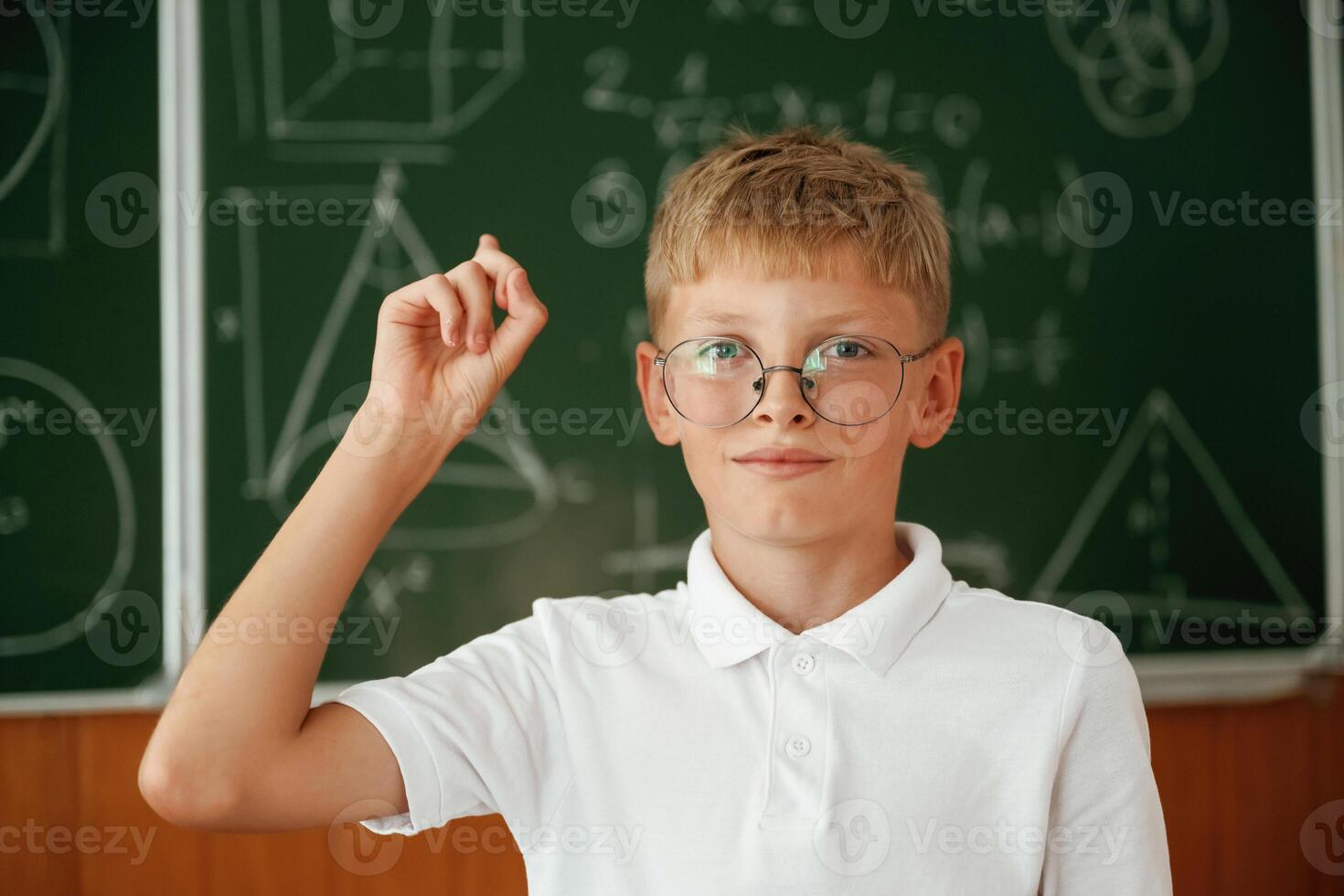 Kid in glasses ,,,is standing in ,the c,lassroom against the blackboard with graphs, hand up photo