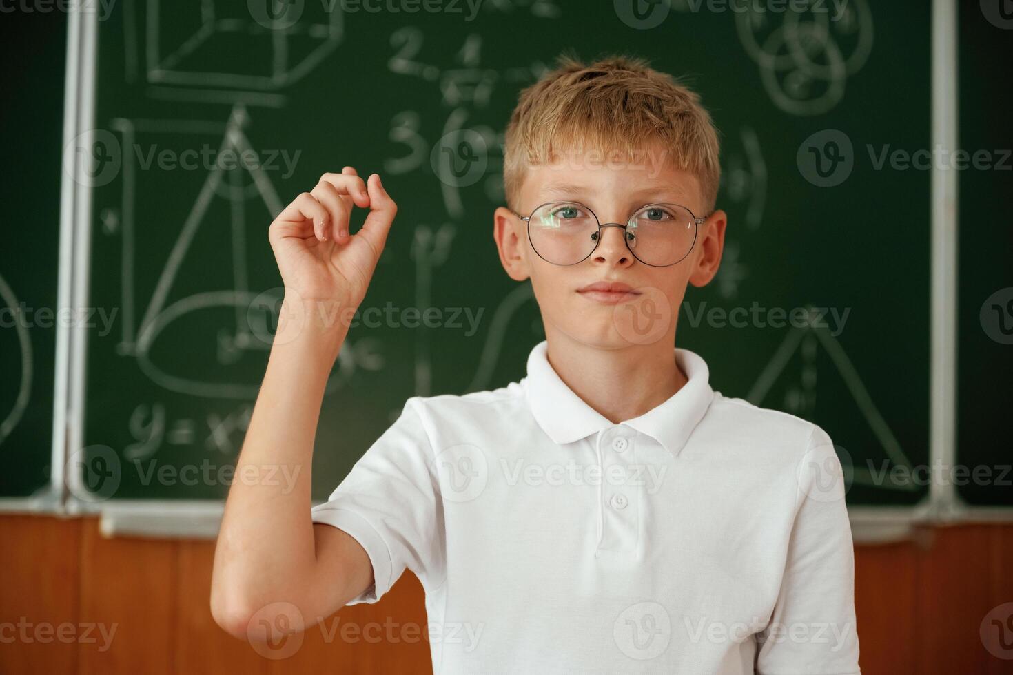 Kid ,in glasses is standing in the classroom against the blackboard with graphs, hand up photo