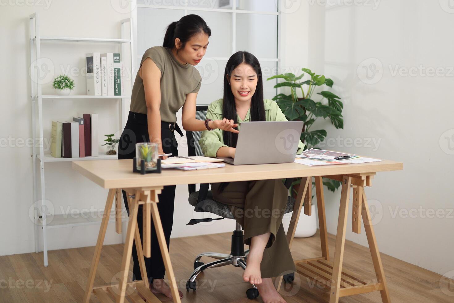Two female employees are working together at a desk, discussing documents while looking at a laptop screen. Business ideas and strategies photo
