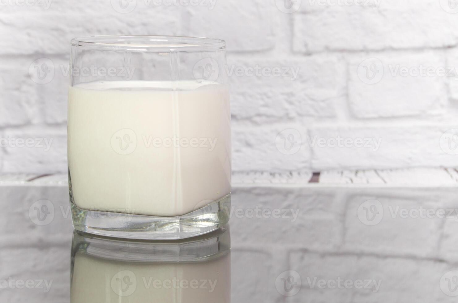 Milk in a clear glass cup on the table, reflection, against the background of a white brick wall photo