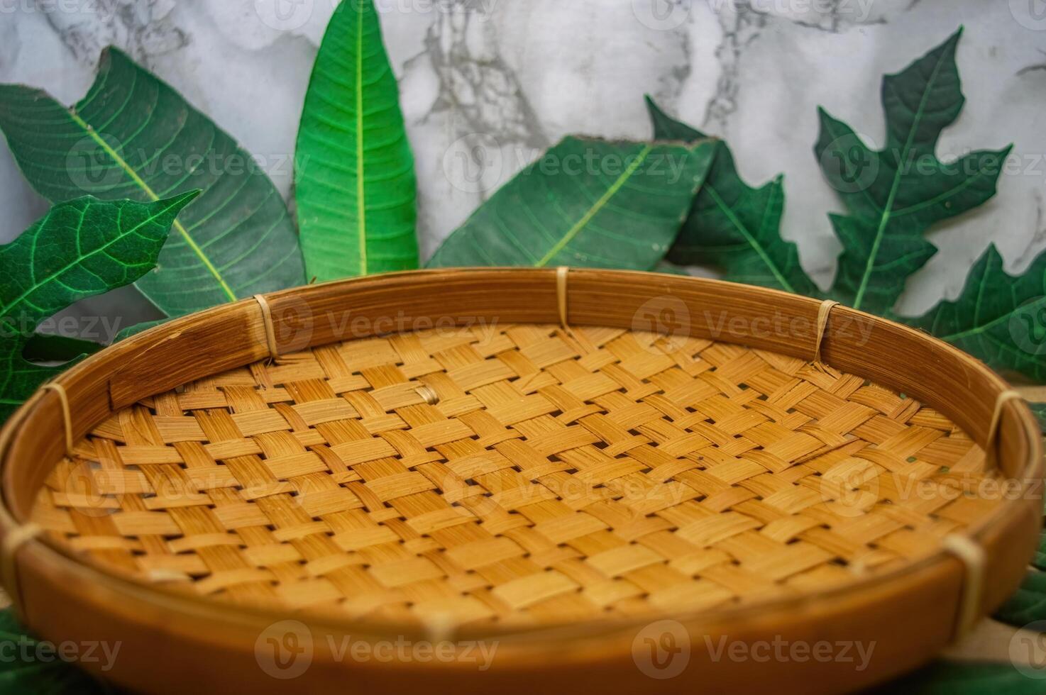 A beautifully crafted round tray of woven bamboo, showcasing intricate patterns and warm tones, set against a backdrop of lush green leaves, perfect for natural product presentations or rustic theme photo