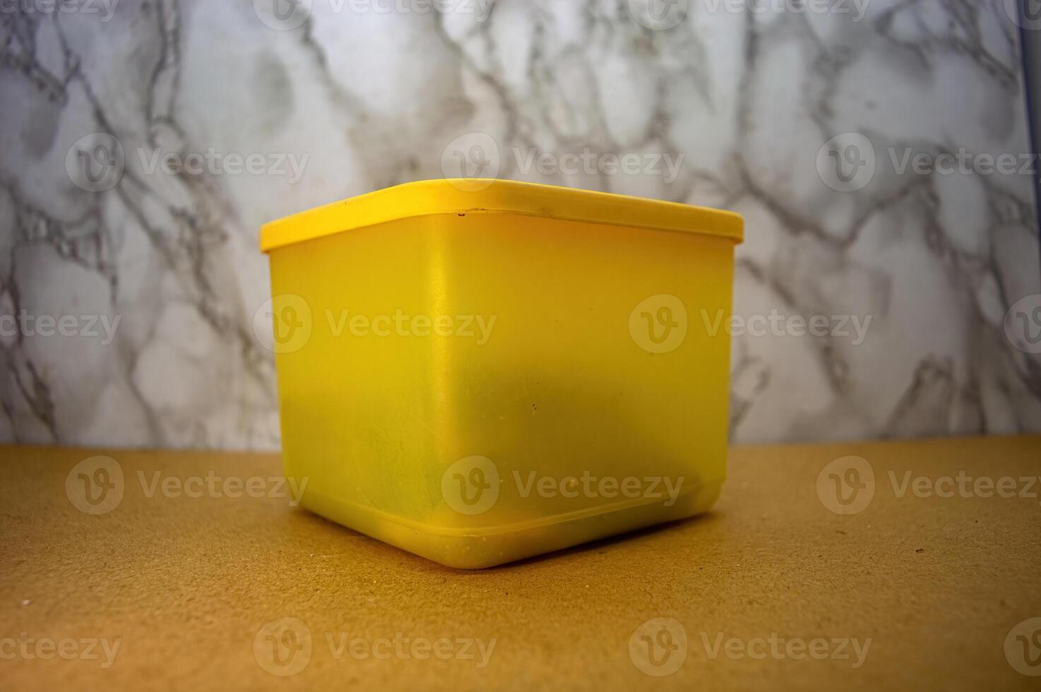 A plain yellow plastic container, subtly translucent, standing on a light brown surface against a blurred marble background, perfect for showcasing storage solutions or a minimalist aesthetic photo