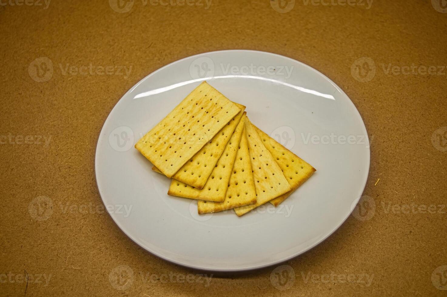 A stack of crisp, rectangular crackers on a white plate, set against a warm brown background. Ideal for snack, food, or appetizer themes photo