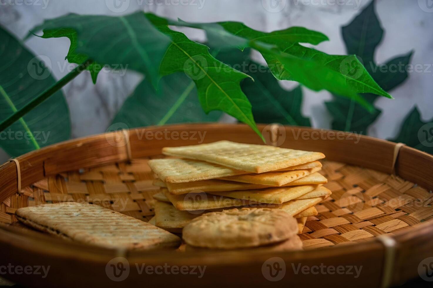 A close up view of golden biscuits and crackers neatly arranged in a rustic woven basket, framed by vibrant green foliage, perfect for a cozy snack or light refreshment photo