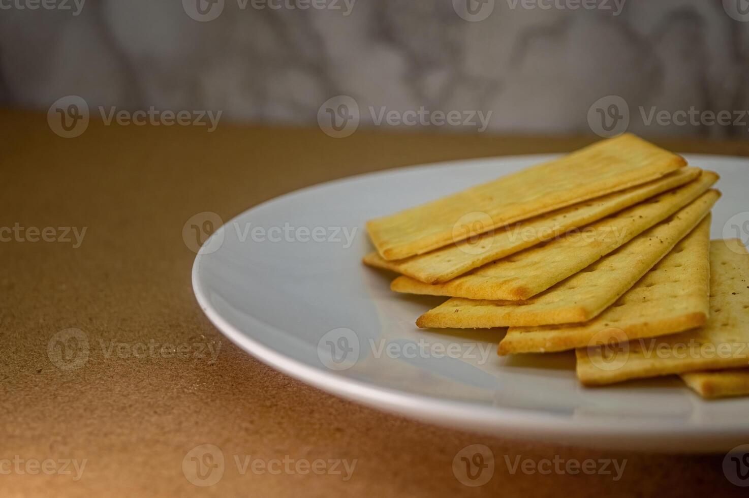 A close up of a stack of golden, rectangular crackers on a white plate, set against a brown surface and subtle textured background. Ideal for food content photo