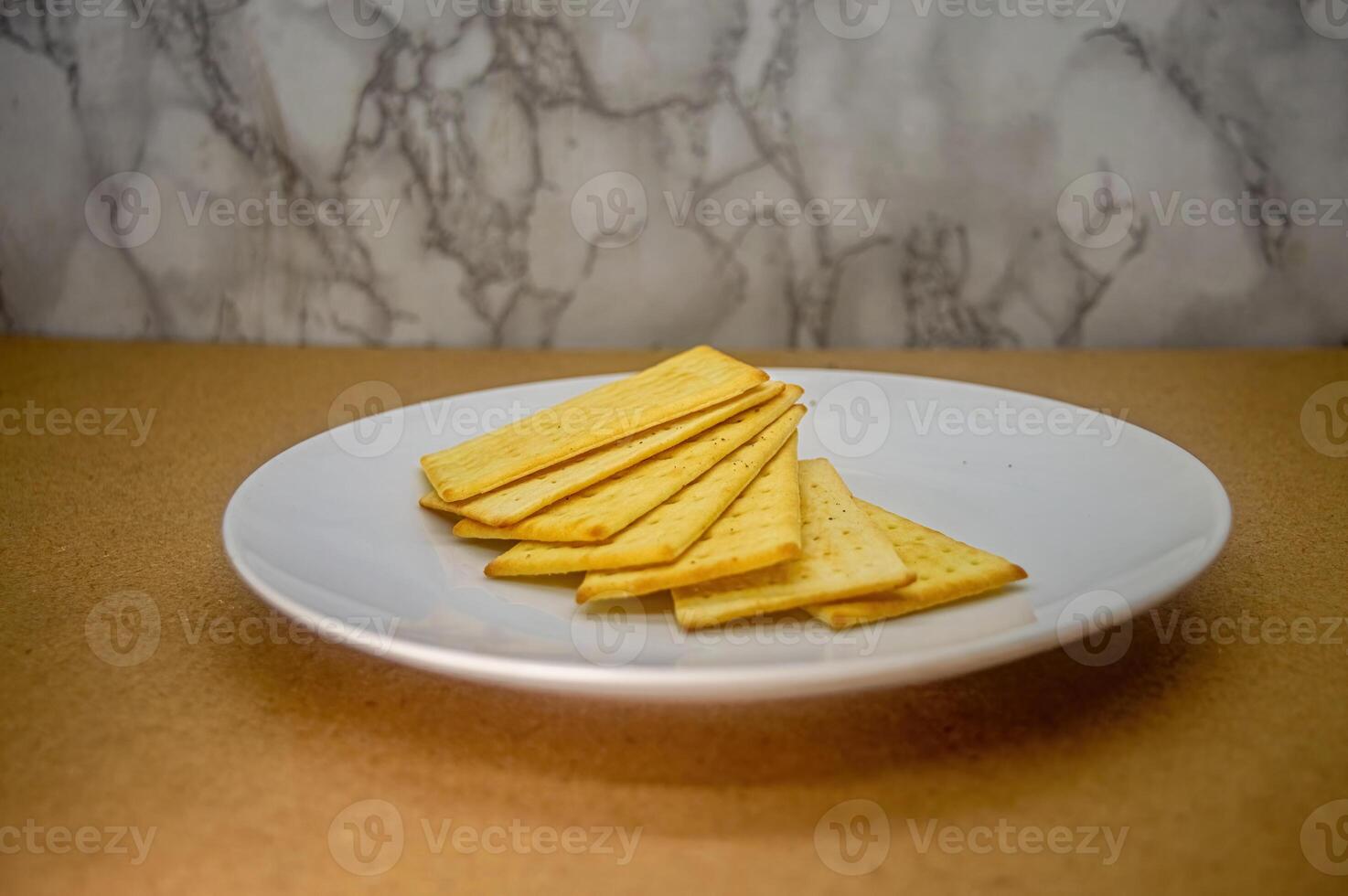 A modest pile of golden, rectangular crackers arranged on a pristine white plate, set against a simple brown surface and textured wall. Ideal for food content photo