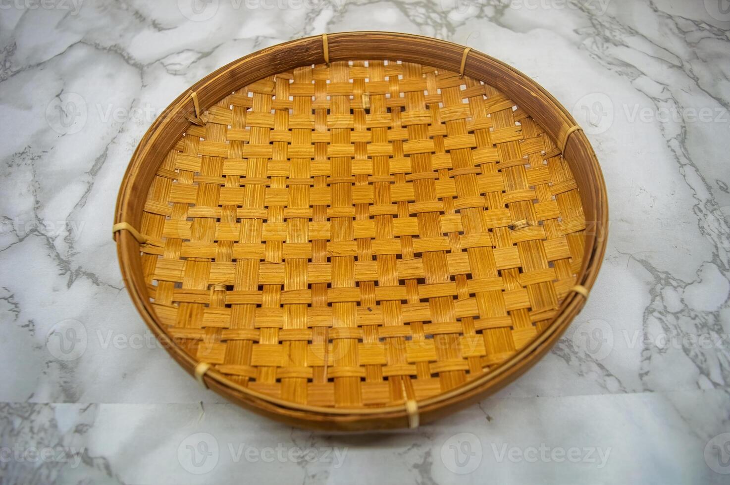 A traditional round woven bamboo tray or tampah, beautifully textured and resting on a white and gray marble surface, providing a versatile backdrop for natural or cultural themes photo