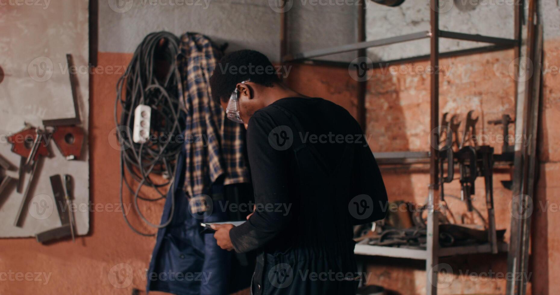 Blacksmith in goggles uses a digital tablet to search for ideas for work, sitting down at his desk and starting to sketch, working in a forge among a variety of tools. in 4k, red komodo photo
