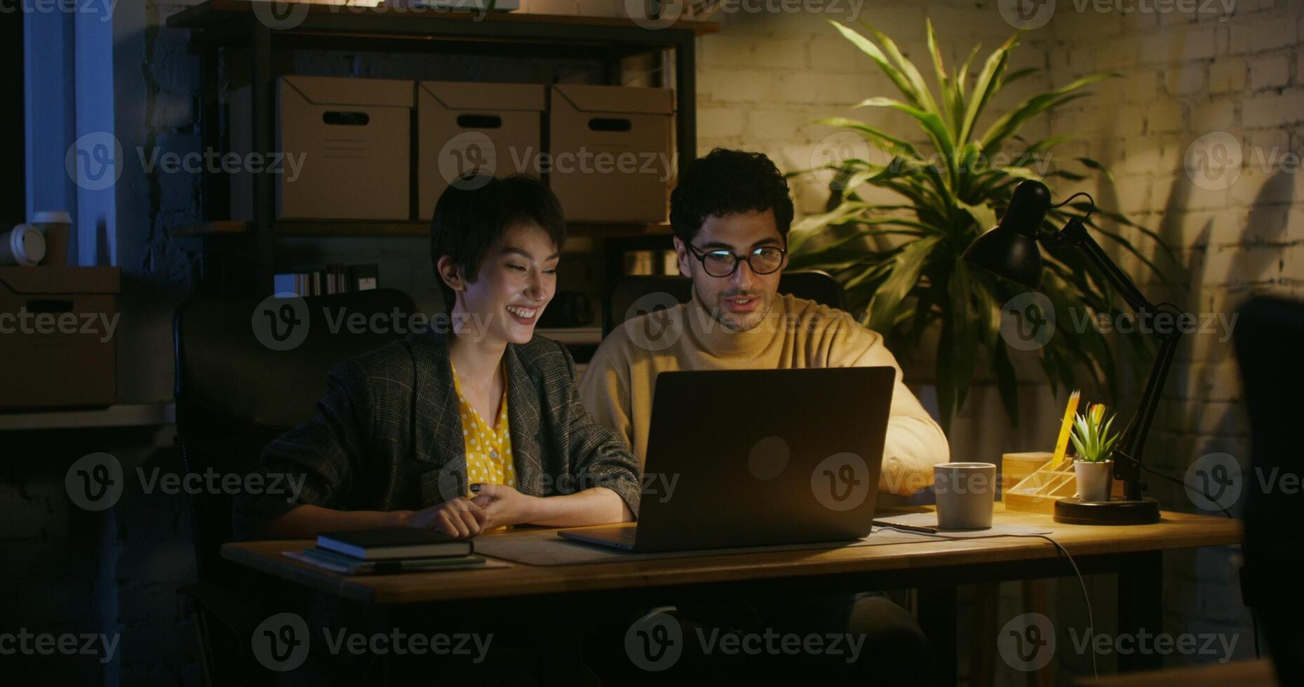 Young handsome man showing young woman to a colleague something on the laptop screen and explaining. They both laugh happily while looking at the screen while sitting at the desk in the office. photo