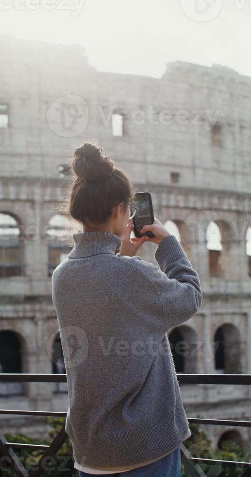 A young beautiful lady taking photos of the sights of Rome on a mobile phone, standing on an observation deck near the Coliseum. in 4k, red komodo, vertical
