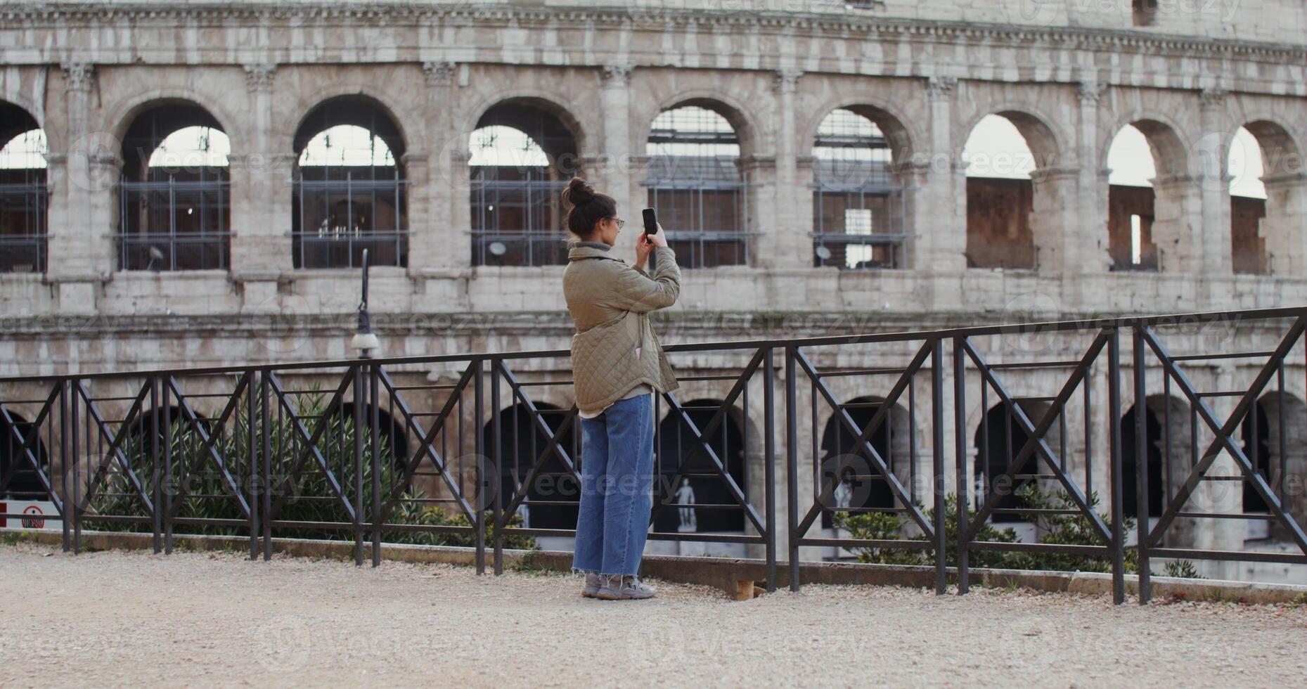 Warmly dressed woman of European appearance takes photographs of the sights of Rome on a mobile phone, standing on an observation deck near the Coliseum. in 4k, red komodo. photo
