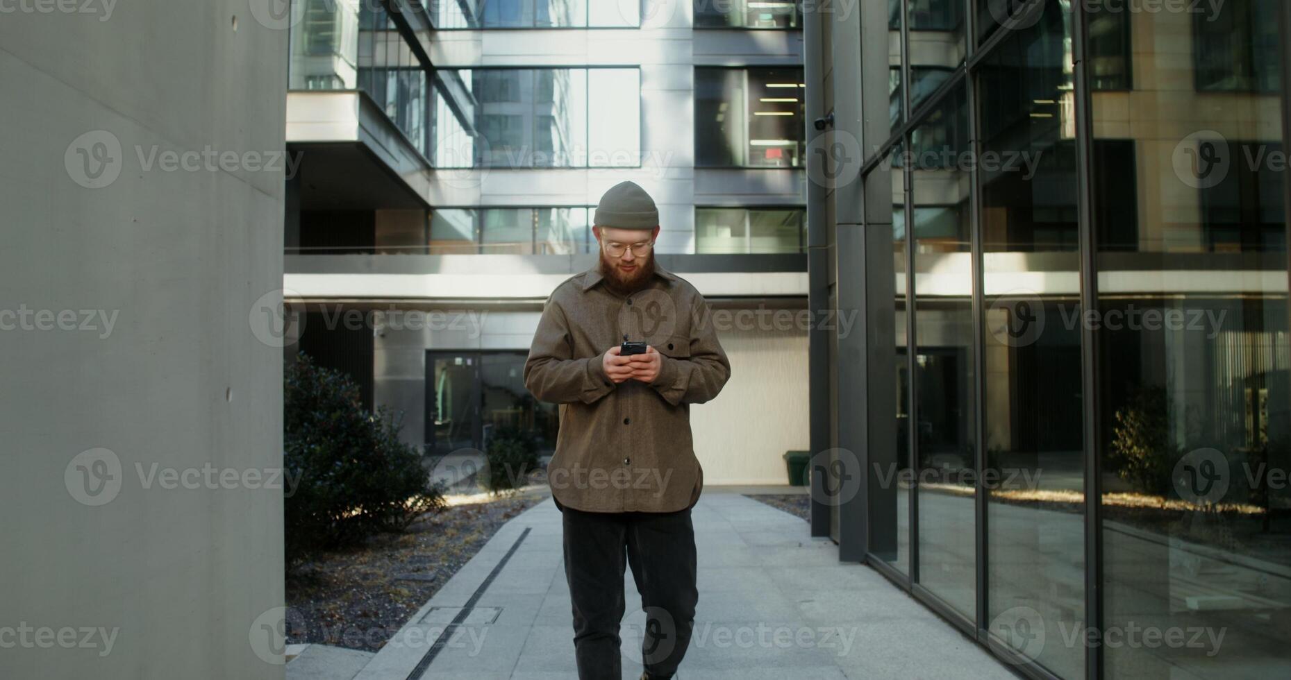 Young man with a beard, dressed in casual clothes and a knitted hat, is typing on a mobile phone while walking in the courtyard of a modern glass building. in 4k, red komodo photo