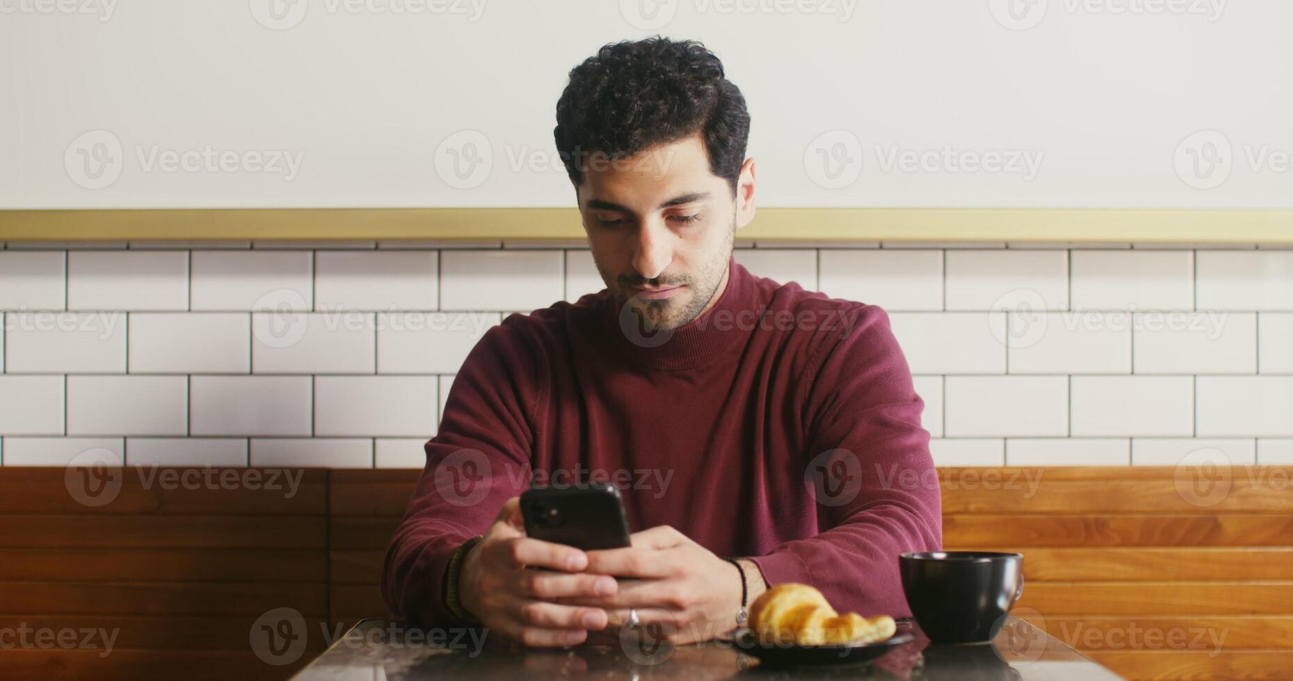 Young Georgian man with stubble on his face, sitting at small table in a cafe, drinking coffee and typing on the phone, having breakfast alone photo