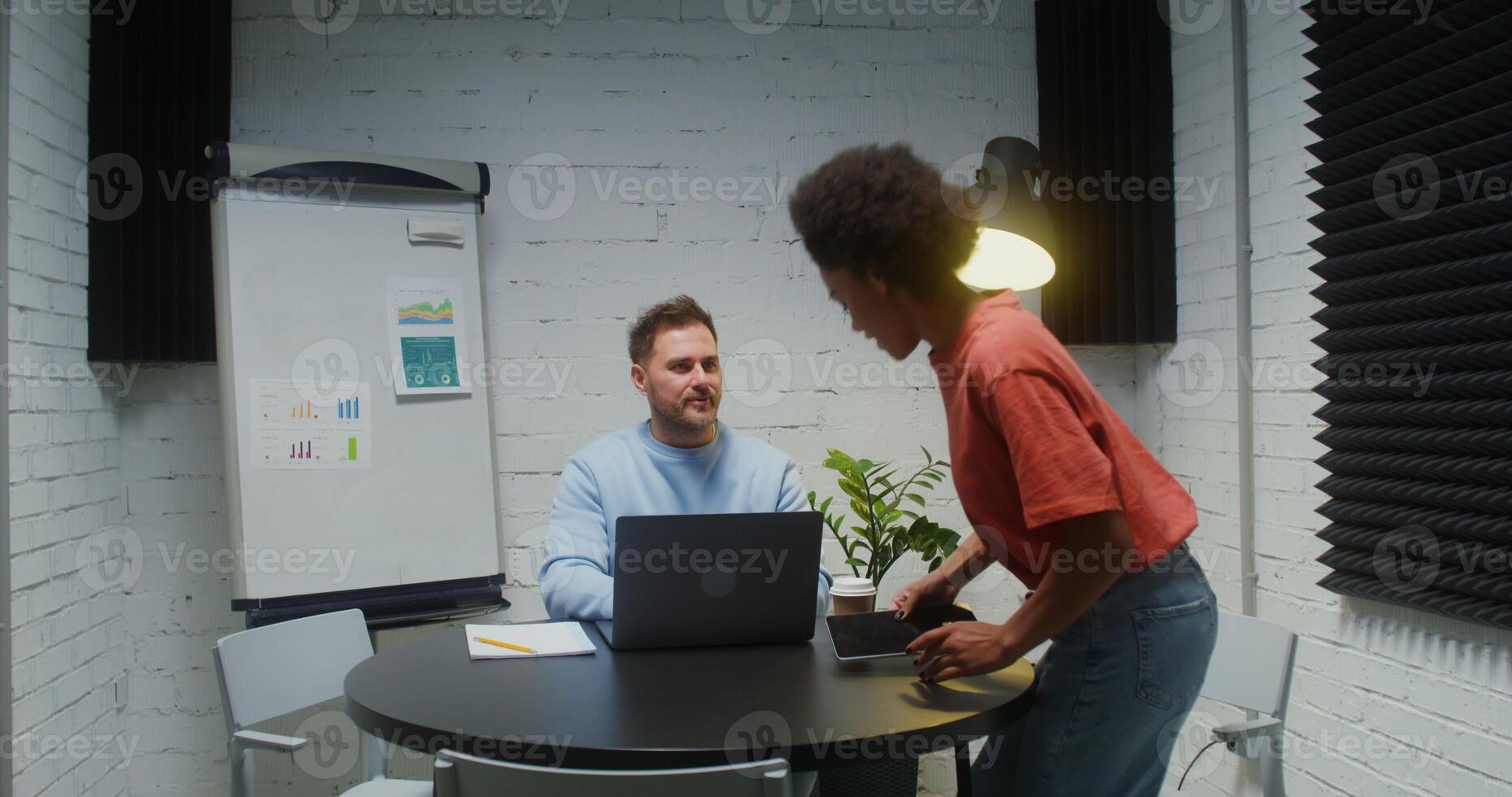 A man of European appearance working at a laptop, welcomes a young woman who comes into his office and starts a dialogue with her, praising her project photo