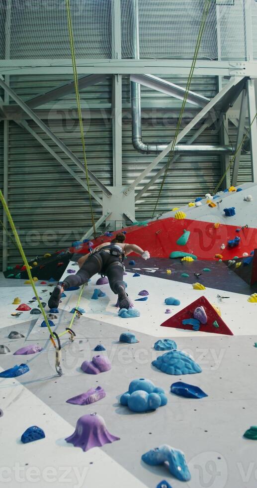 Athlete of European appearance treats her hands with talcum powder, lowering them into a bag attached to a belt behind her, while climbing to the top of the climbing wall, vertical photo