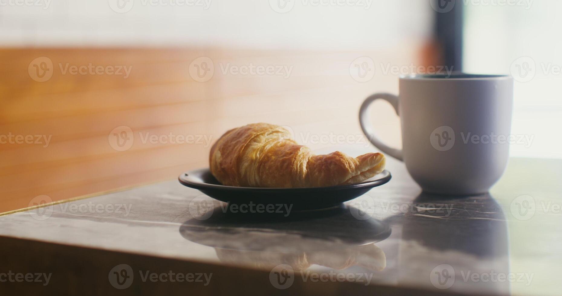 A large white mug with steaming hot coffee and black plate with a croissant stand on a small table in a cafe, ready for lunch photo