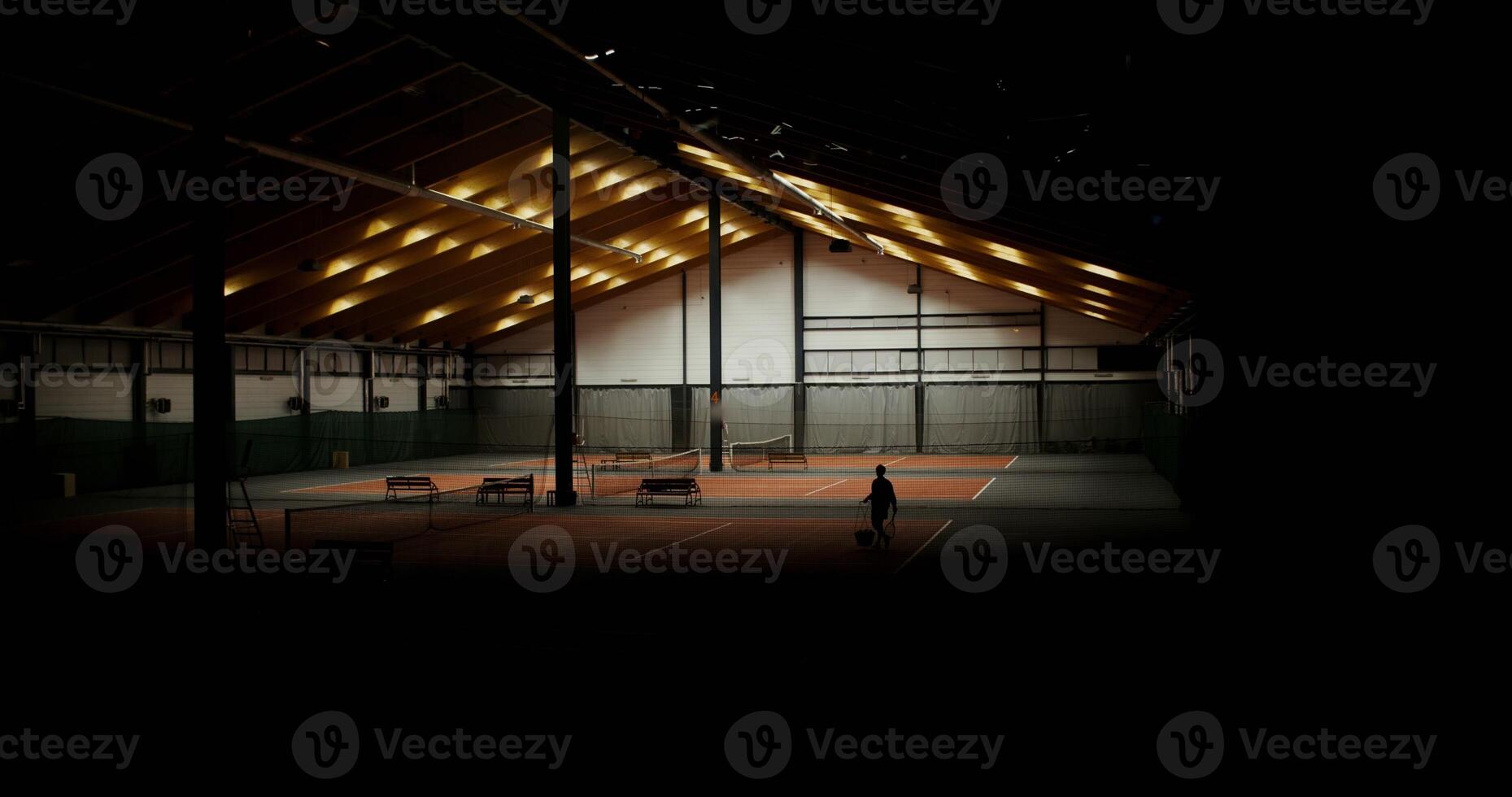 Large indoor sports hall with several tennis courts, lights are turned on and a man is seen walking towards the bench with a racket and a basket full of tennis balls in his hands. photo