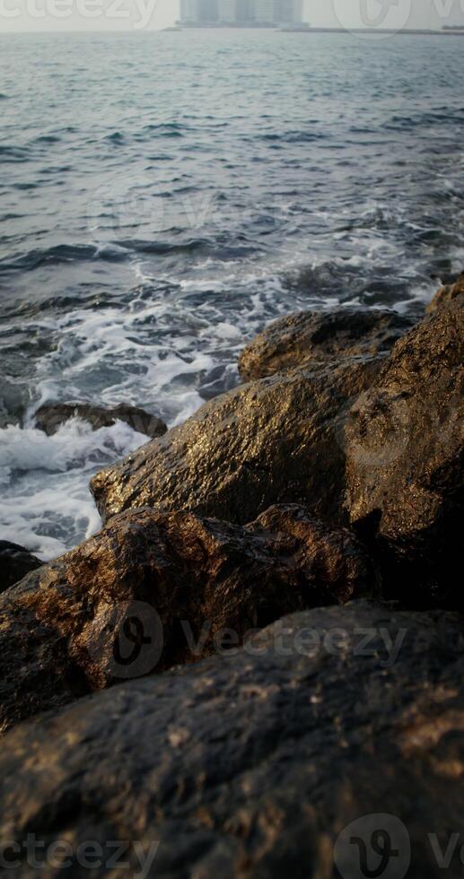 Sea waves hit large rocks on coast, foaming and splashing as they wash over the boulders. The hotel complex is visible on the peninsula in the distance, vertical . 4k , red komodo photo