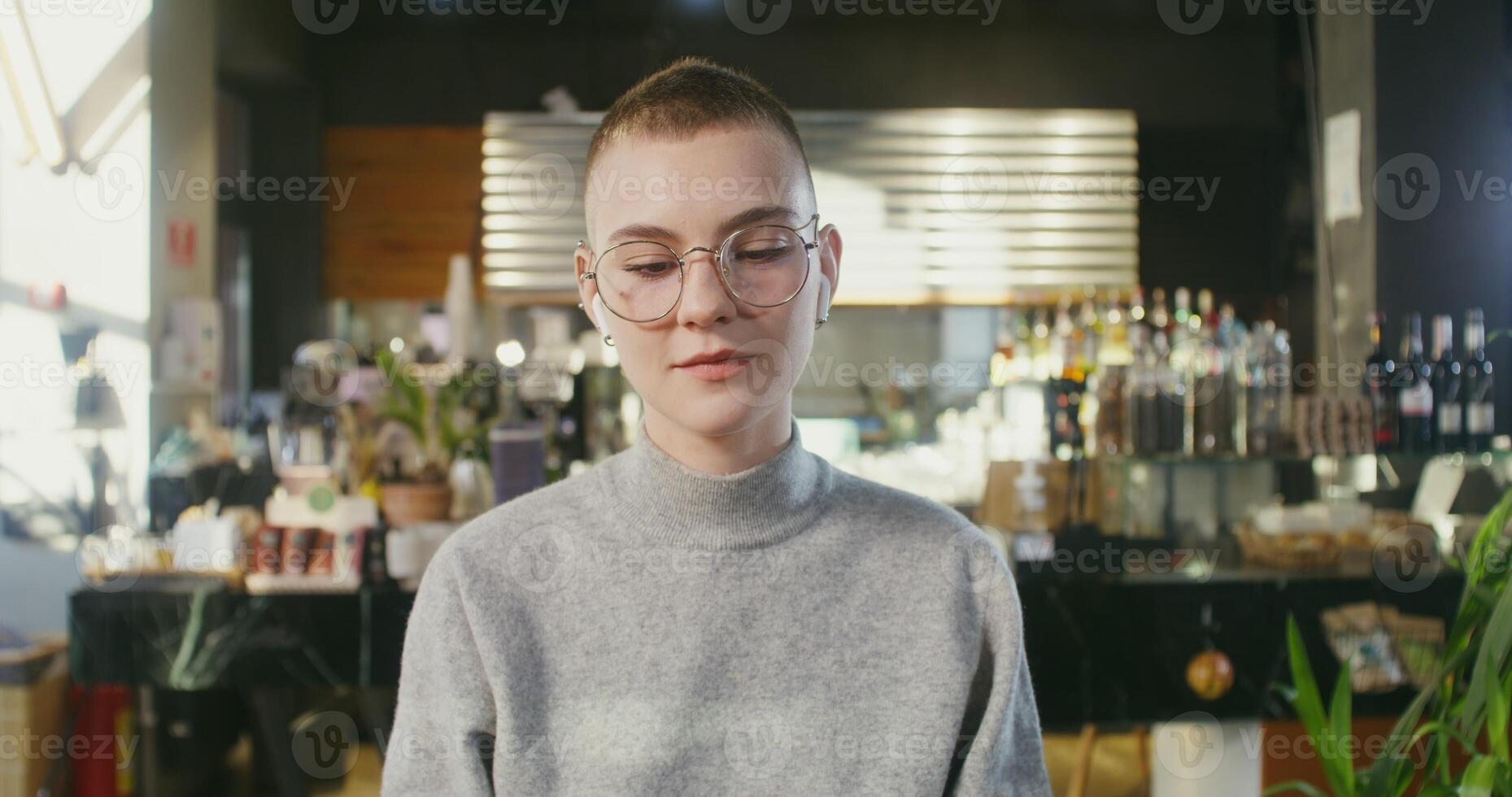 Young woman with hedgehog of hair on her head, wearing glasses inserts wireless headphones into her ears, opens the lid of a laptop and answers a call while sitting in a cozy coffee shop photo