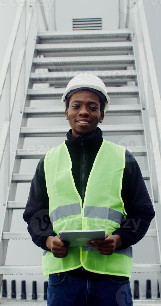 A young man in white helmet and a green vest looks at the camera with a smile while standing near the entrance to the wind turbine with a tablet in his hands, vertical . in 4k, red komodo photo