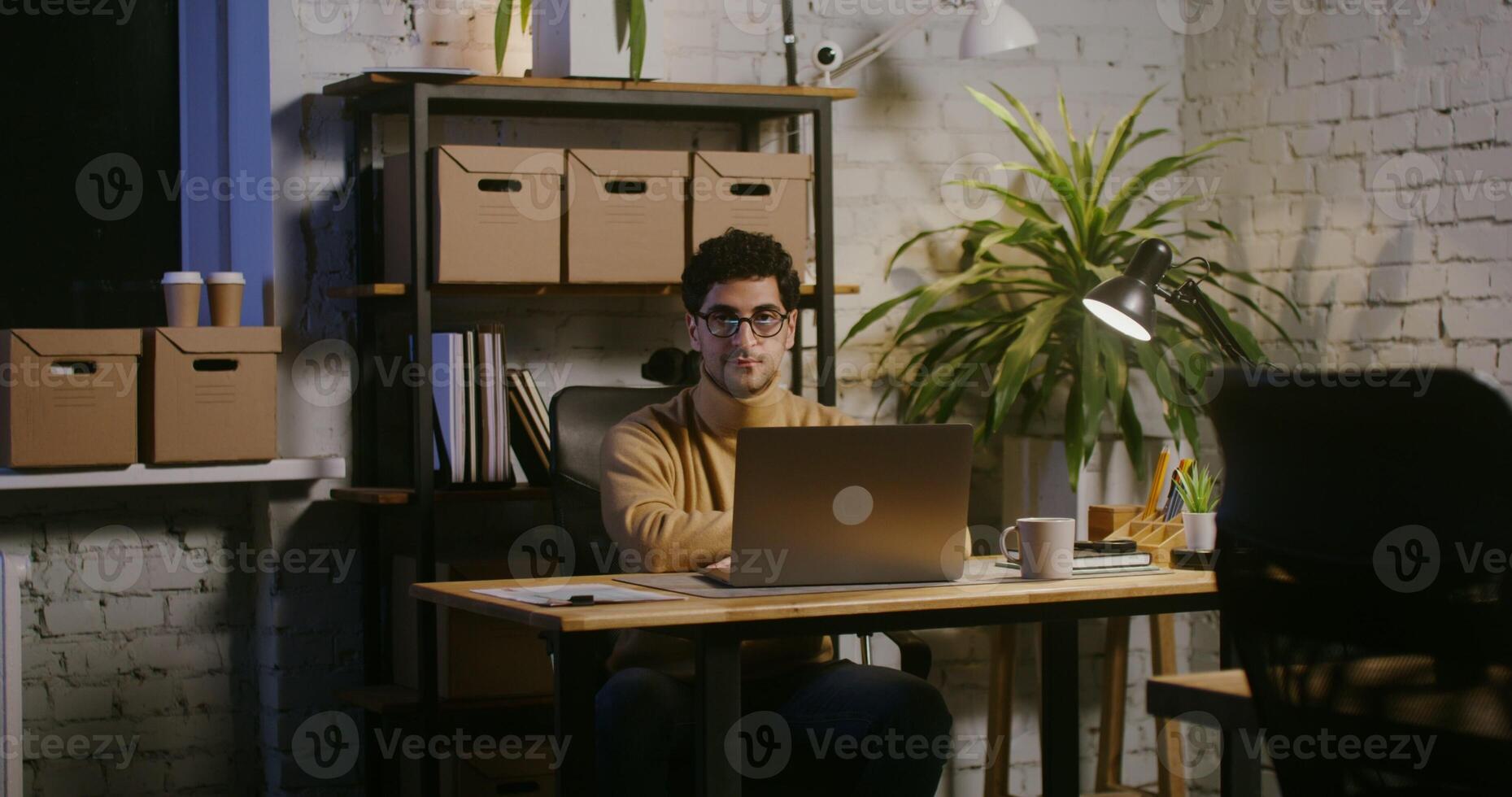 Young man with stubble on his face looking straight into the camera and then taking off his glasses, while sitting at the table in front of an open laptop in a modern stylish office photo