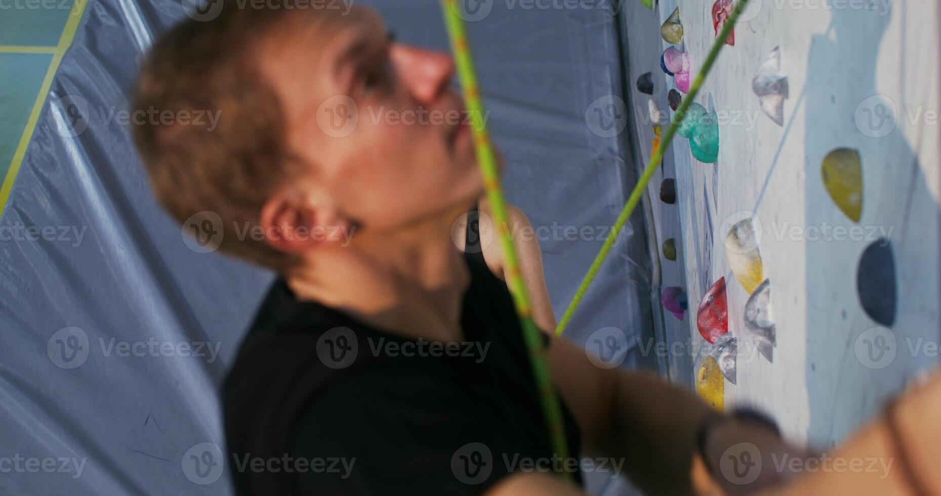 A young woman watches man gravely as he climbs the climbing wall and holds a safety rope while standing at the foot of the wall. Indoor gym climbing training photo
