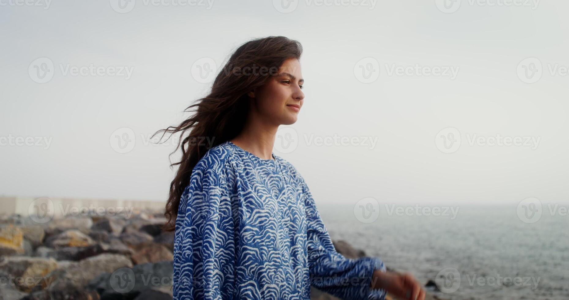 A young beautiful woman straightens her dark curly hair, standing on the sea coast photo