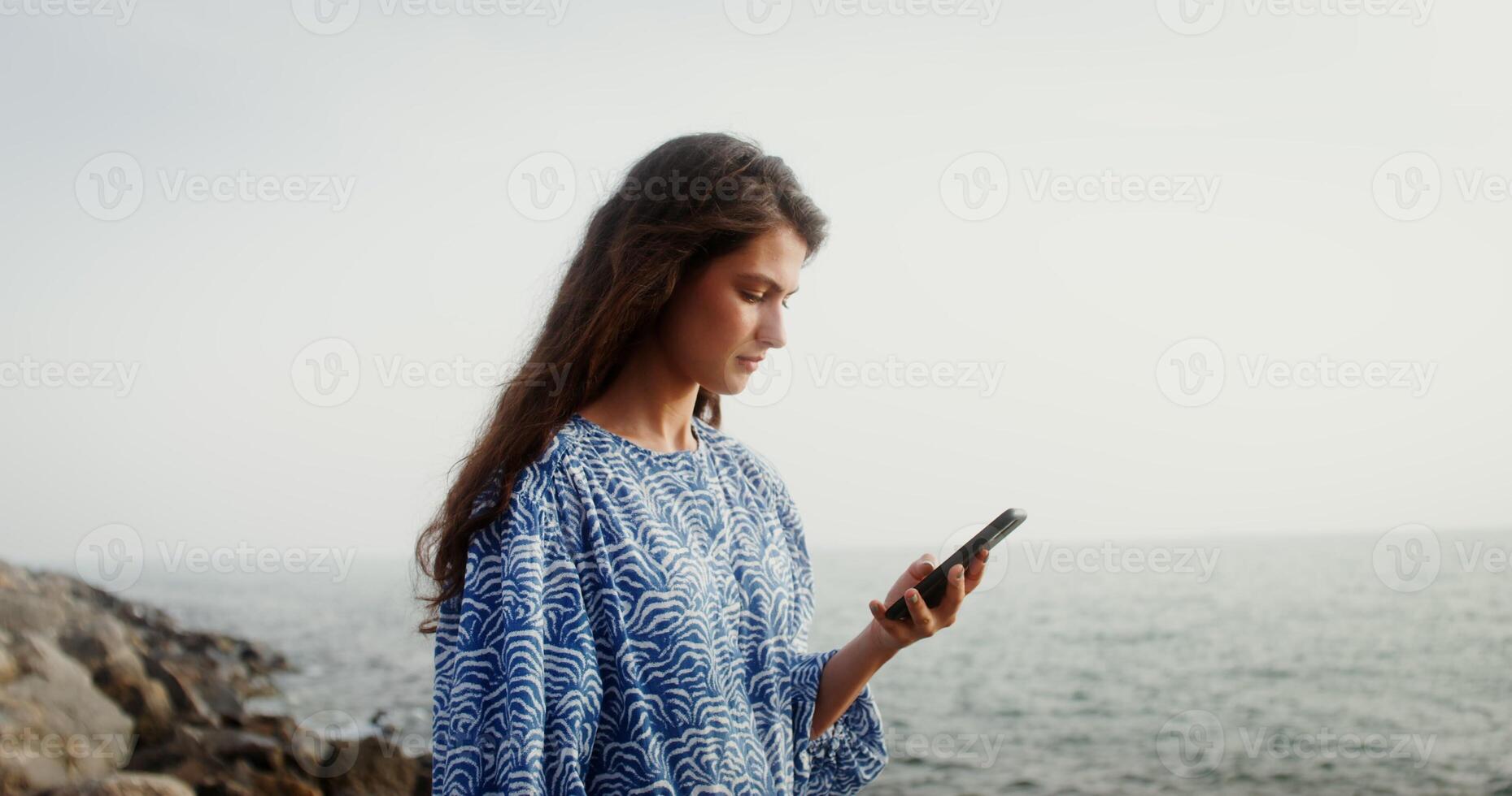 Young beautiful woman using mobile phone while standing on a rocky sea shore. Social networks, correspondence on a smartphone outdoors. 4k , red komodo photo