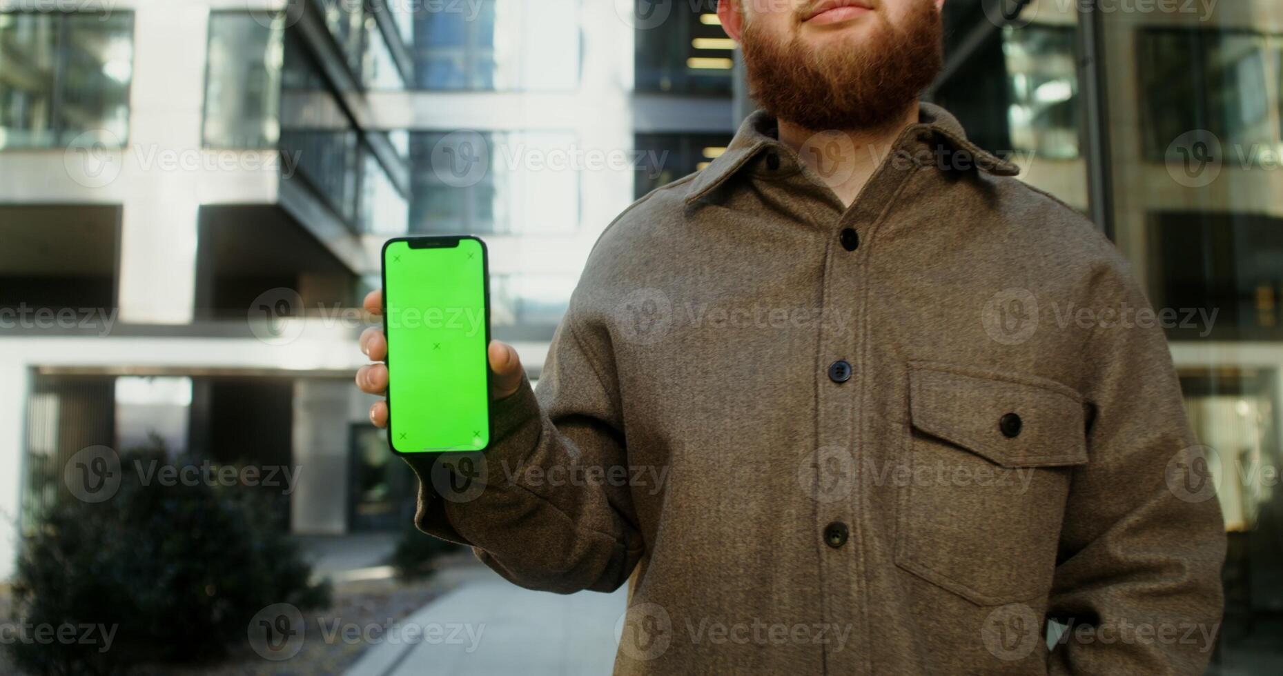 Mobile phone face forward in the hands of young man dressed in casual clothes, standing in the business center of the city. Copy space, green screen, chroma key, mockup. in 4k, red komodo photo