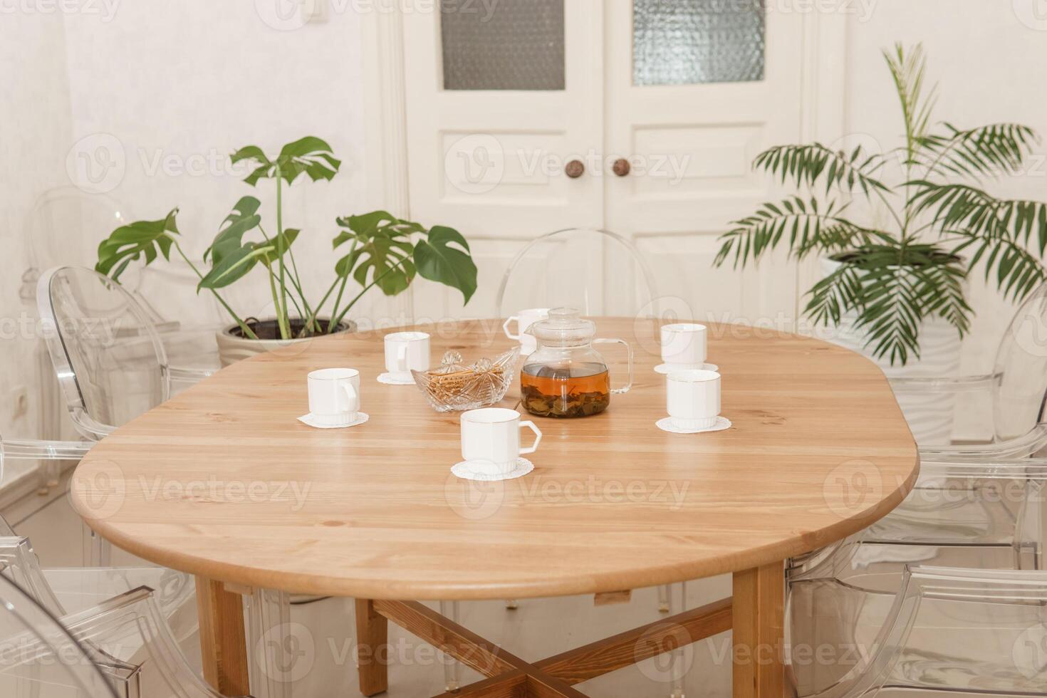 Cozy dining setting featuring round wooden table surrounded by transparent chairs, complemented by lush green potted plants, creating warm and inviting interior atmosphere. photo