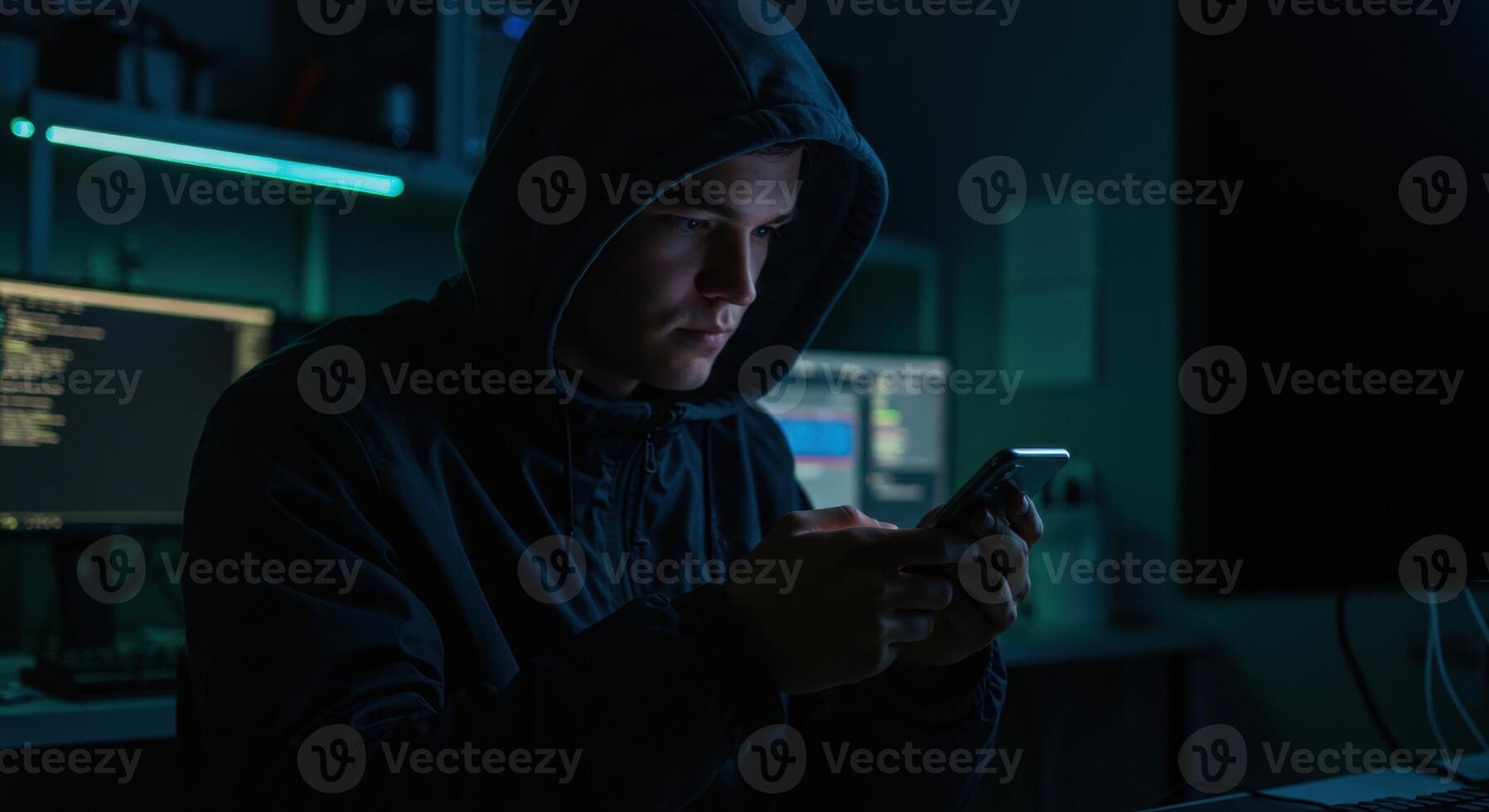 Young person in a dark hoodie using a smartphone in a dimly lit room with computer screens. photo