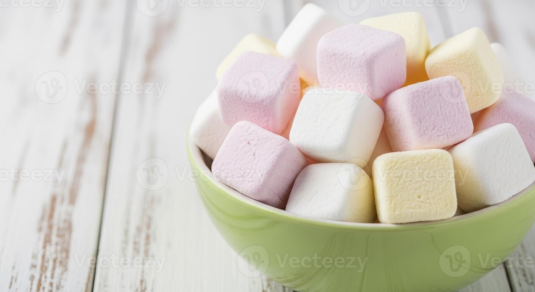 Soft powdery white pink and yellow cube marshmallows in a green bowl on a rustic wooden surface photo